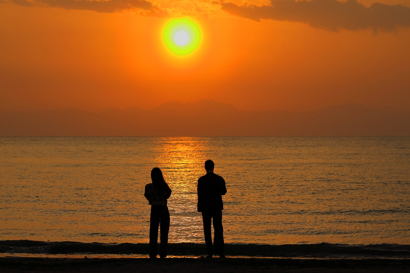 オレンジ色に染まる夕暮れの逗子海岸、波打ち際で夕日を眺めたたずむカップルの後ろ姿