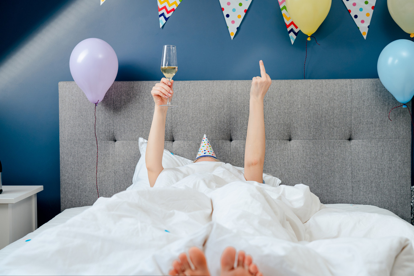 Covered by blanket woman lying on bed with arms raised, holding glass of champagne and showing middle finger, fuck off sign. Passive Birthday plans. Time for yourself. Holiday at home. Happy