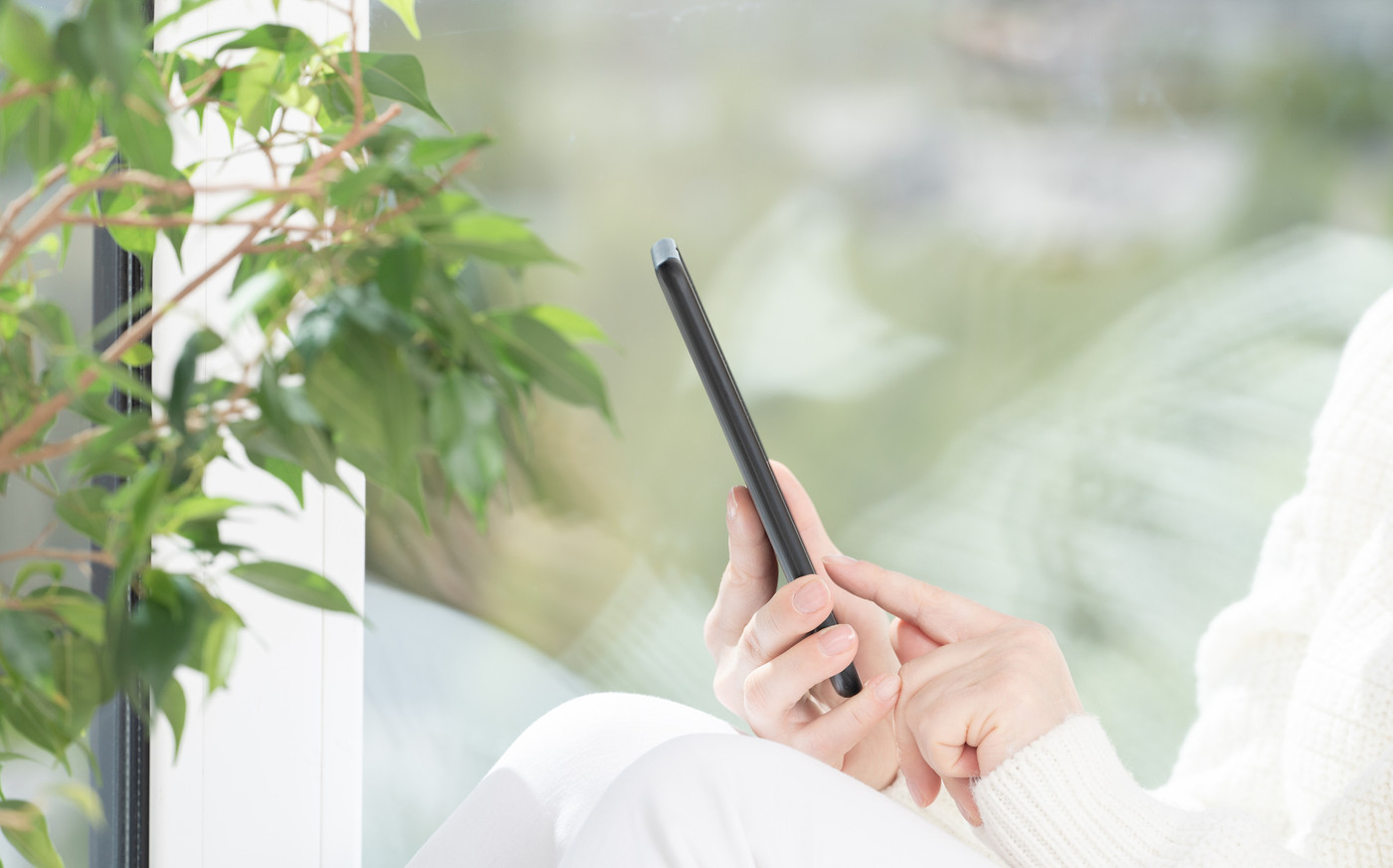 Girl in white clothes sits by window and uses cell phone, looks at smartphone, reads, shop, communicates with friends in messenger. reading text message on cell telephone at home.
