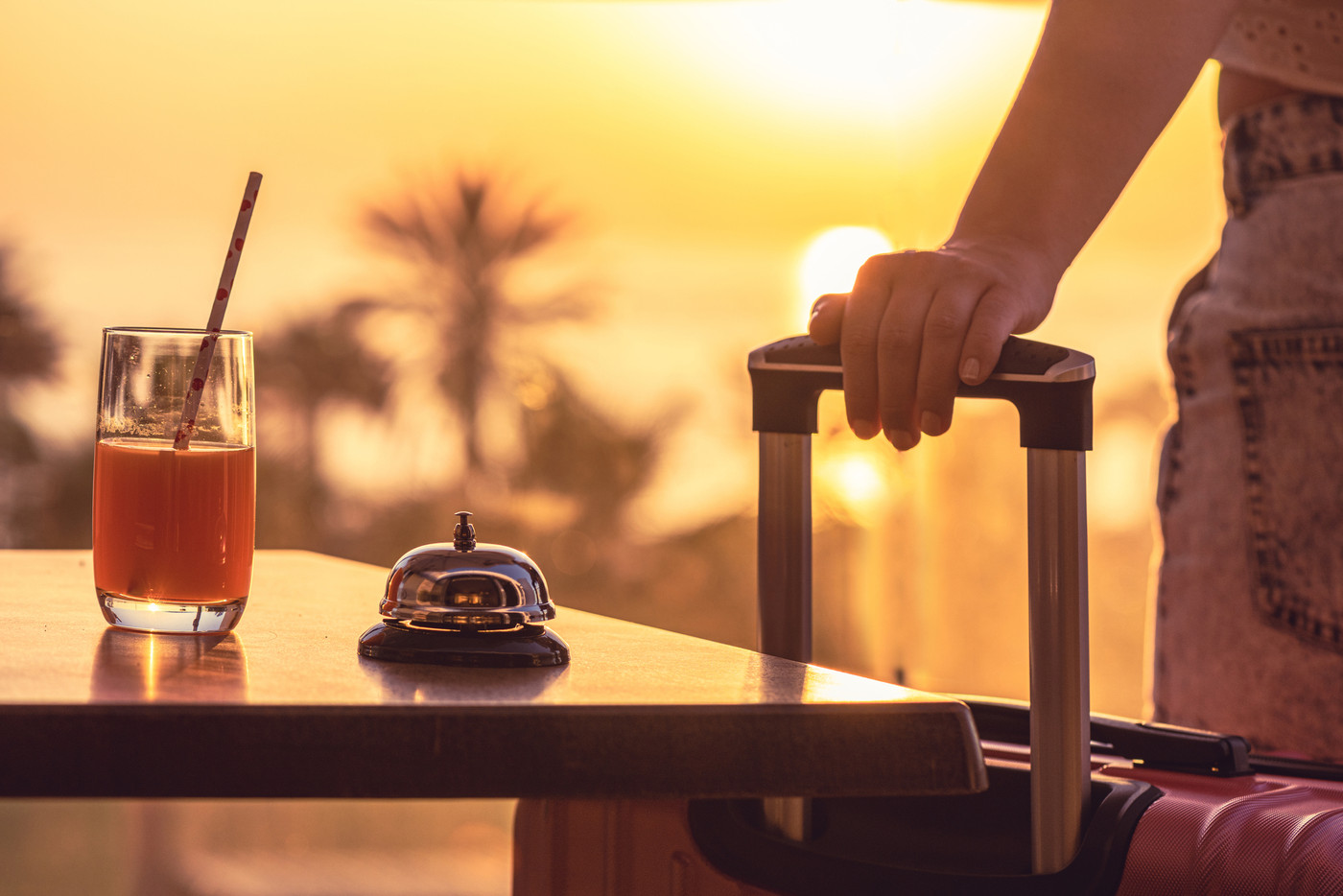 Woman with suitcase ringing hotel service bell with welcome drink and sea and palm tree view on sunset. Travel concept. 24-hour beach hotel front desk. Late check-out.