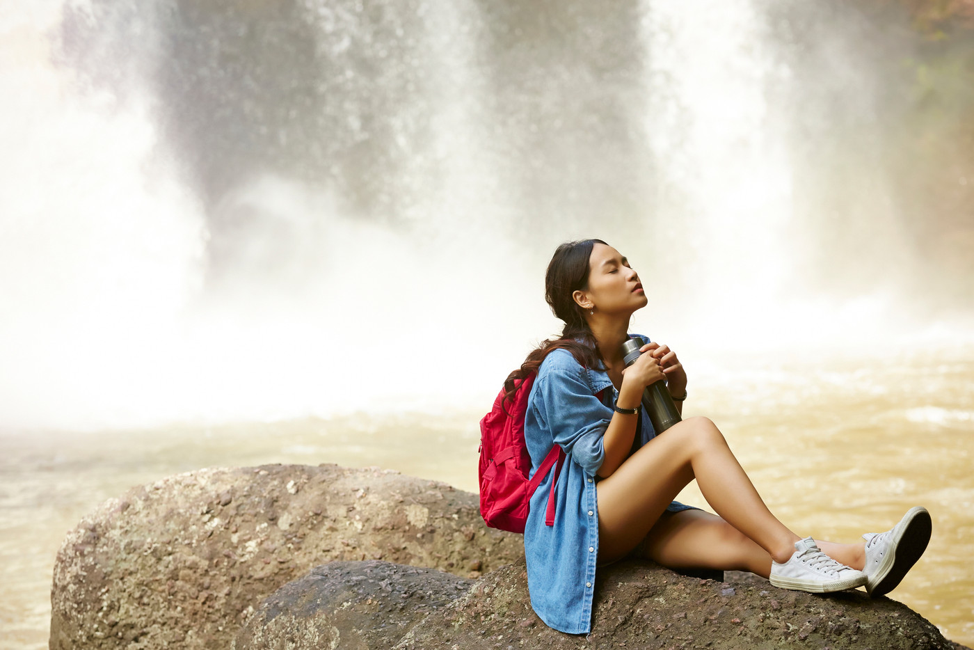 the girl with a red backpack is resting on the rock near a waterfall