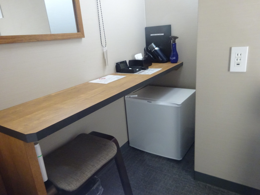 デスクと椅子。奥に冷蔵庫、ドライヤー、消臭スプレーあり。