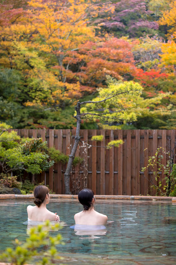 Back view of two young women relaxing in an open-air bath with autumn leaves in the background