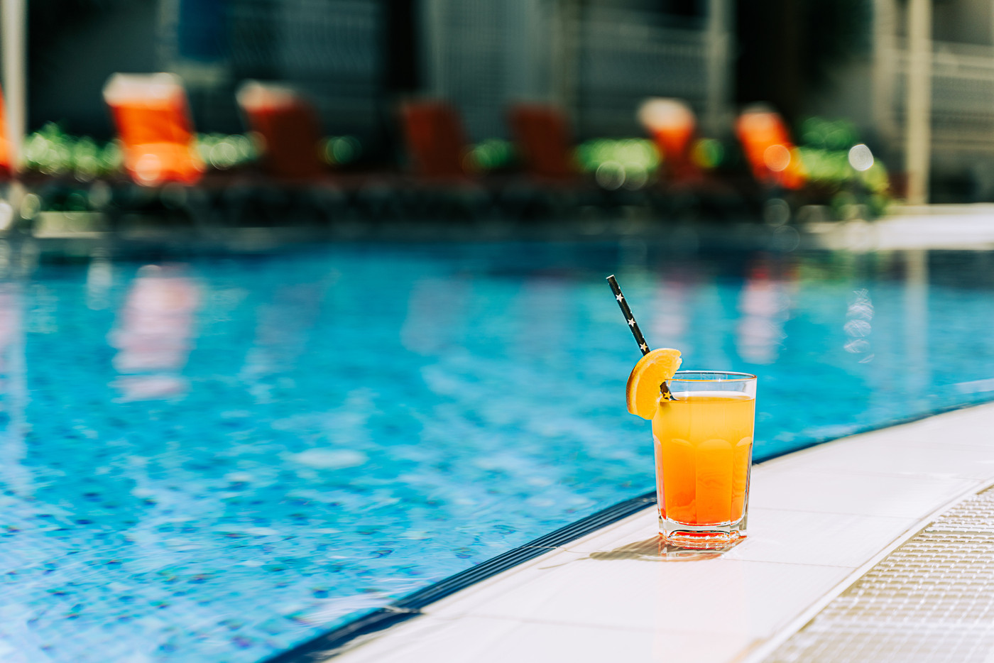 Tropical sparkling cocktail by the pool. The shot of glass with orange lemonade fruit cocktail standing near the poolside. Summer alcohol free drink by the hotel pool. Hello summer holiday v
