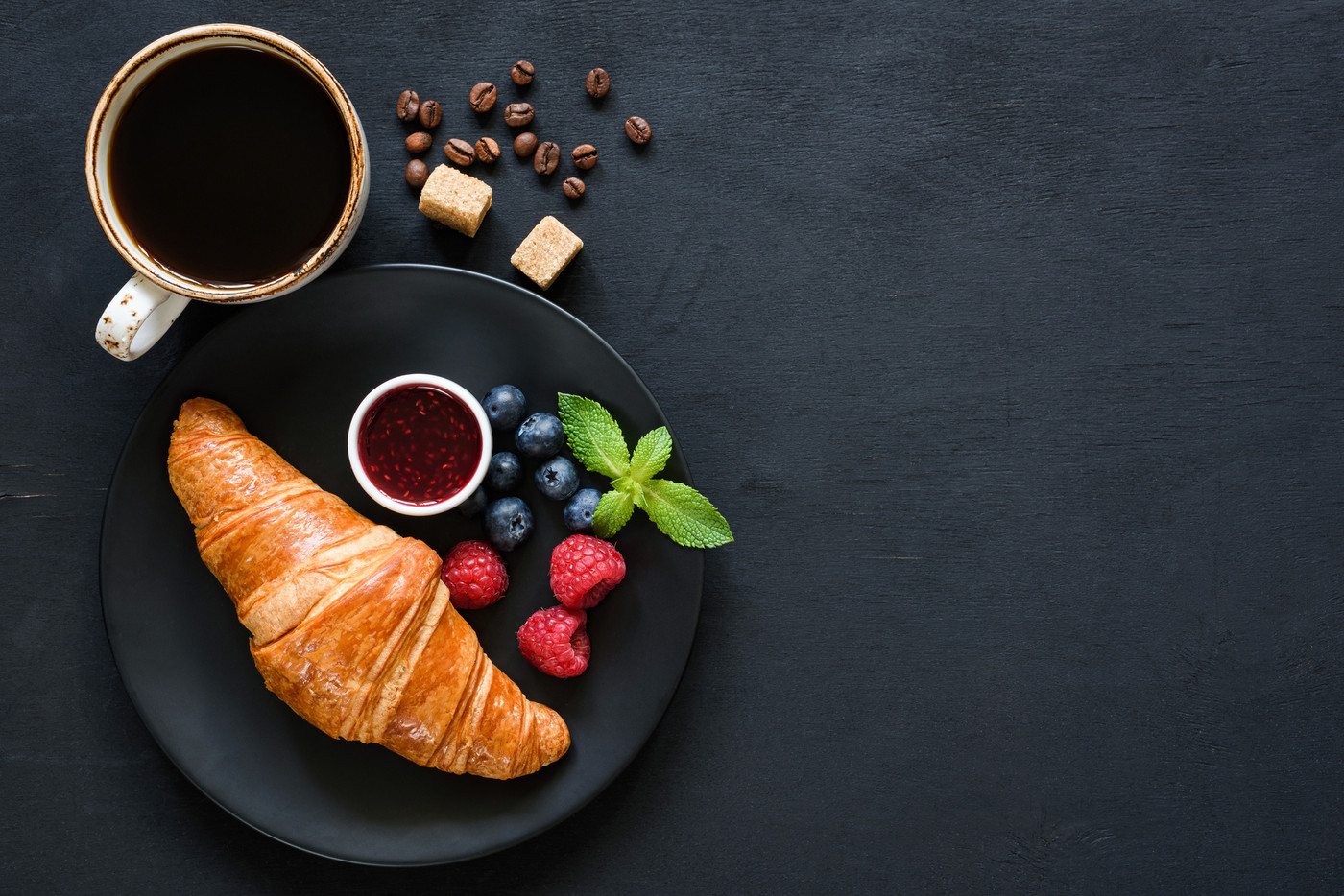 Croissant with berries, jam and cup of black coffee on black background