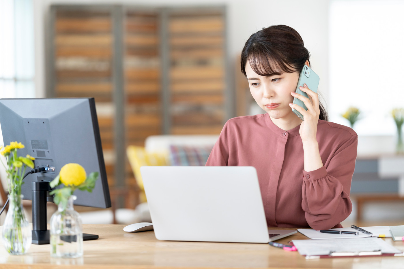 スマートフォンで通話しながらパソコンの画面を見る若い女性