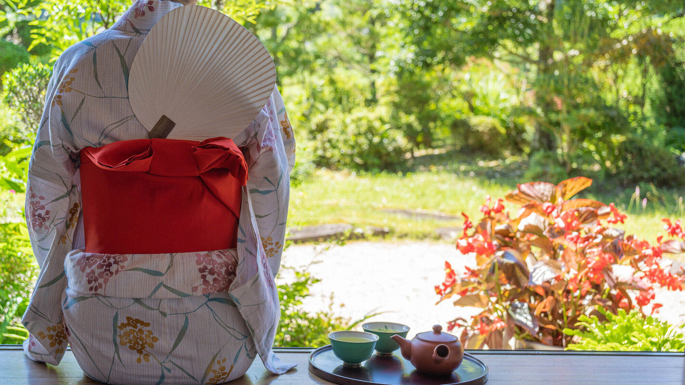 日本の夏のイメージ｜浴衣を着た日本人女性の後ろ姿。縁側で緑茶を飲む。背中にうちわ。
