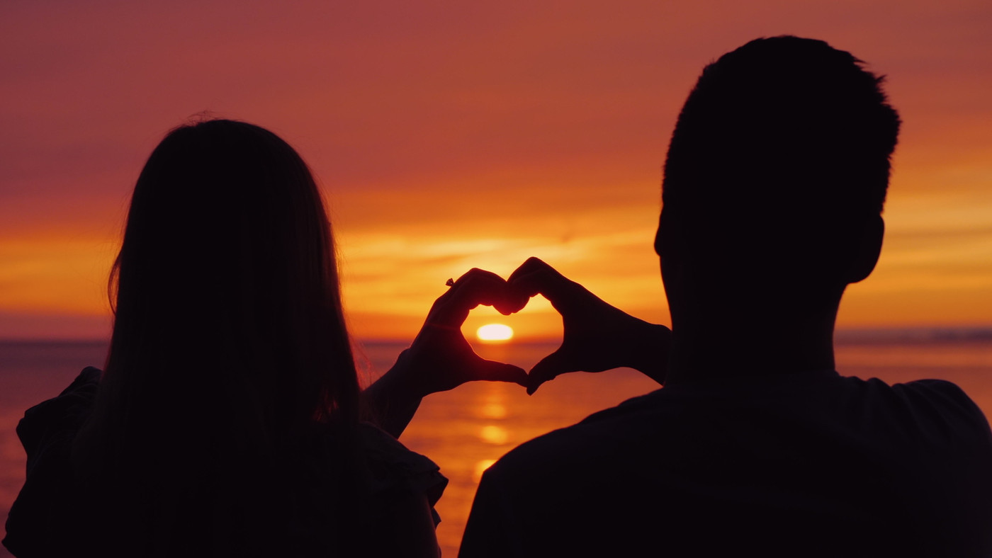 Silhouettes of a young couple, hands show the shape of the heart against the backdrop of the sunset over the sea